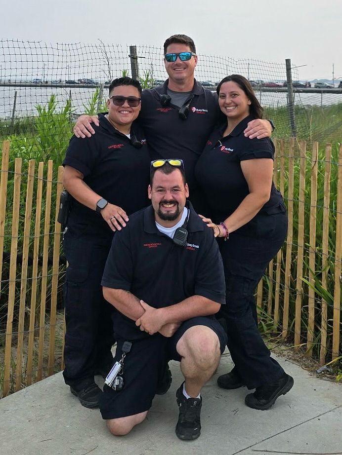 Four people wearing matching black uniforms posing outdoors with one person kneeling in front.