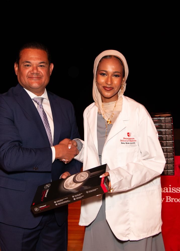 Two individuals at a formal event, one in a suit and the other wearing a white coat and hijab, shaking hands while holding a certificate from the Renaissance School of Medicine.