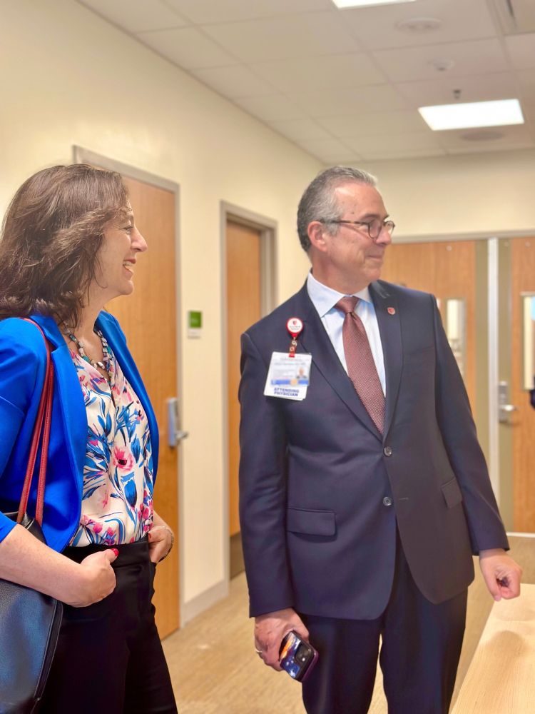 Two individuals, one wearing a blue blazer and floral top, the other in a dark suit with a name badge, smiling and standing in a hallway.