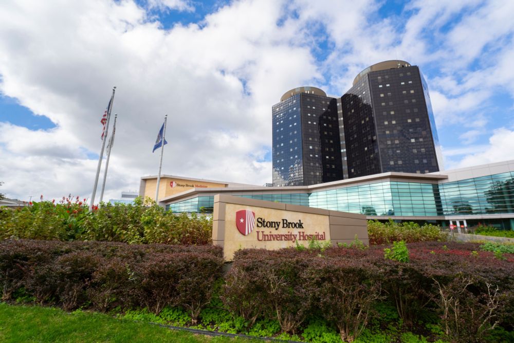 Exterior view of Stony Brook University Hospital with its distinctive round, multi-story towers under a cloudy sky. The hospital's sign is prominently displayed in the foreground.