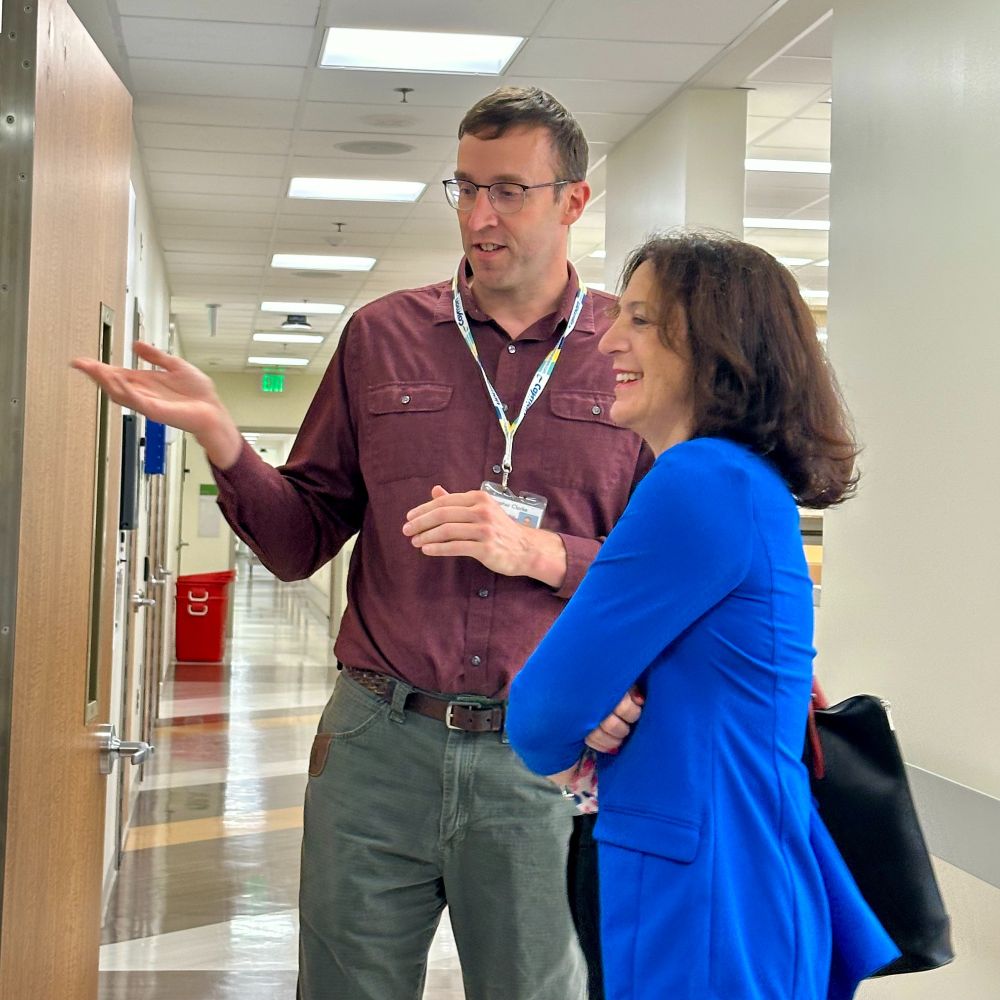 Two professionals conversing in an office hallway, with one person gesturing during the discussion.