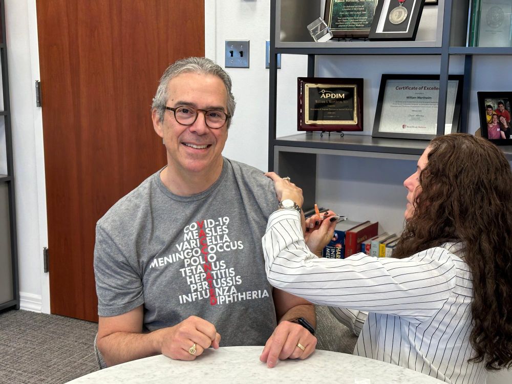Person receiving a vaccine in an office setting, seated across from a medical professional, with a wall of framed certificates in the background.