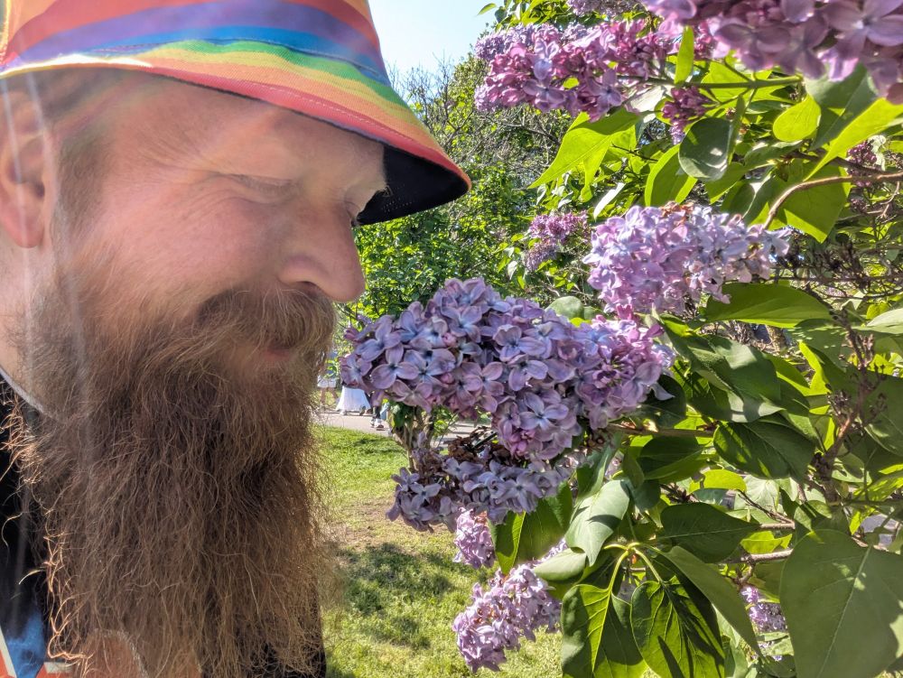 Me wearing a rainbow hat and smelling a light purple lilac flower
