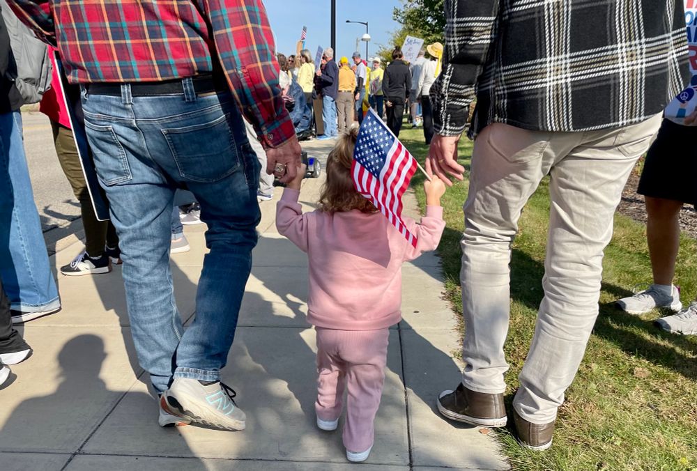 Picture of a young child dressed in pink and holding an American flag, being guided through the crowd slowly by two men in flannels who are pointing out all the chicken and frog costumes to her as she giggles.