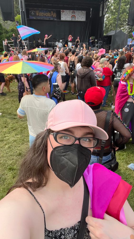 Trans girl wearing a black N95 and a pink cap has the lesbian flag over her shoulder and is standing in a crowd of other gay, trans, nonbinary, and queer people. Mila Jam is performing, and there is a trans flag waving in the crowd