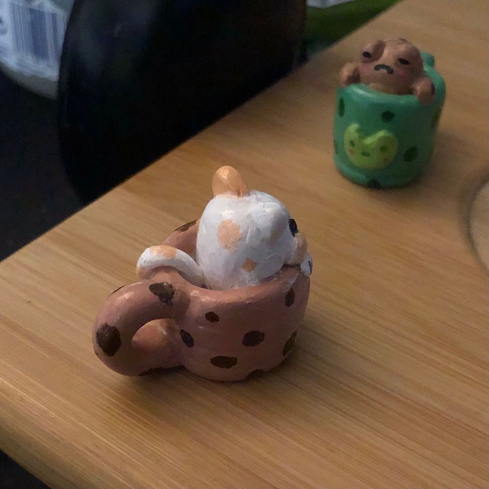 A fat white and orange cat sits in a mug. The mug is pattered like a cookie. 