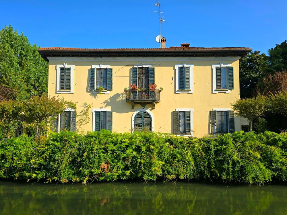 Yellow villa on Naviglio Martesana canal, Milan. The house has a flower balcony and is surrounded by green vegetation.