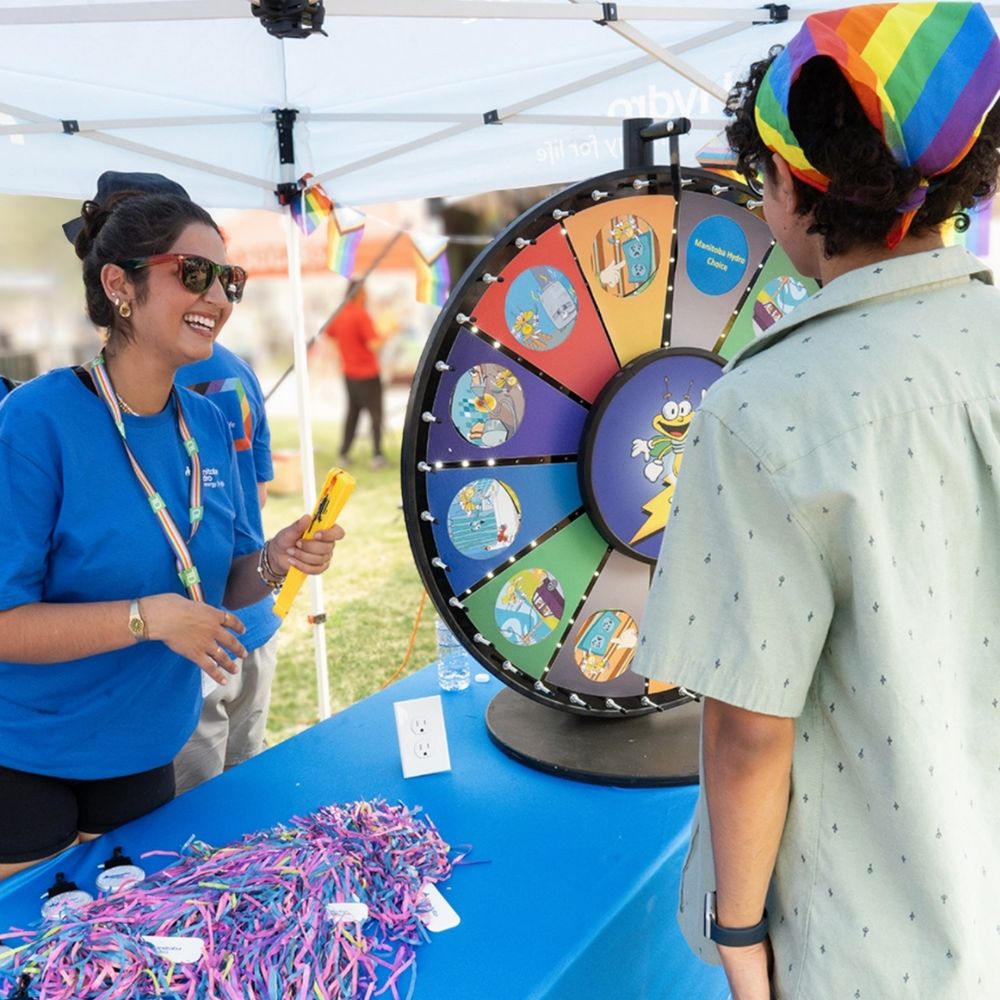 A smiling woman in a blue Manitoba Hydro t-shirt stands behind a booth with prizes. She is talking to a customer next to a spinning prize wheel. 