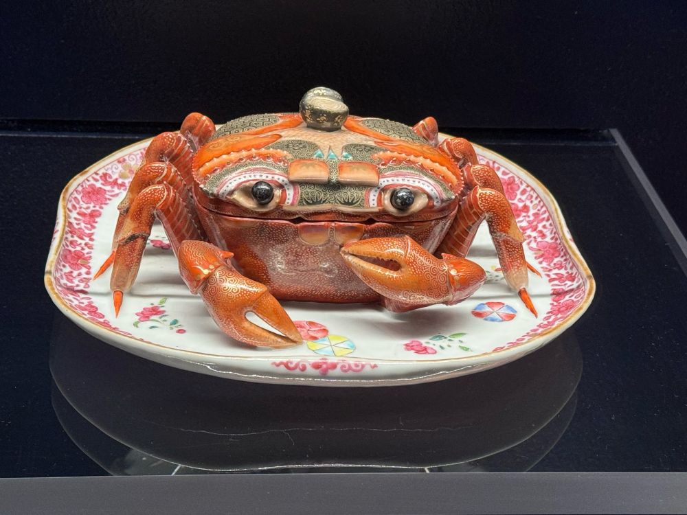 A red soup tureen shaped like crab sitting on a plate. The top half of him lifts off as a lid. He has one claw lifted up towards his chest, and combined with the expression in his eyes, he sees to be saying “who, me? *I’m* a soup tureen?!”