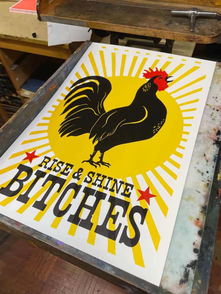 Paper letterpress print of a crowing black & red rooster over a bright yellow sunburst.  Text reads”Rise & Shine Bitches”
