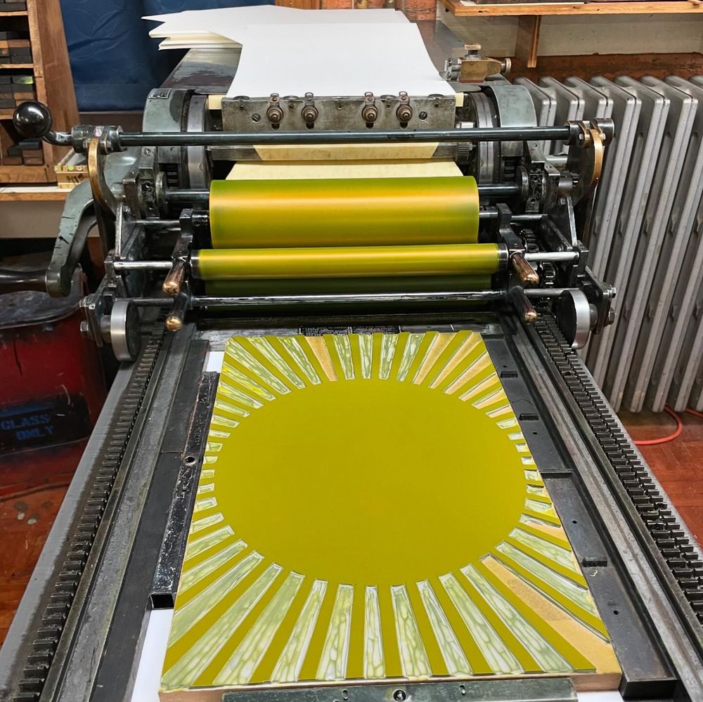 A large carve linoleum block locked in place on the bed of a Vandercook printing press. The rollers and block have yellow ink on them.