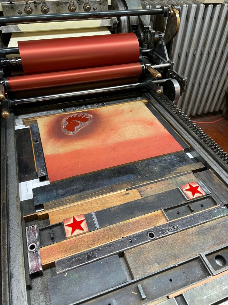 A linoleum cut of the rooster head and 2 woodcut stars locked in place in the bed of a Vander Cook letterpress printing press. The blocks and rollers have red ink on them.