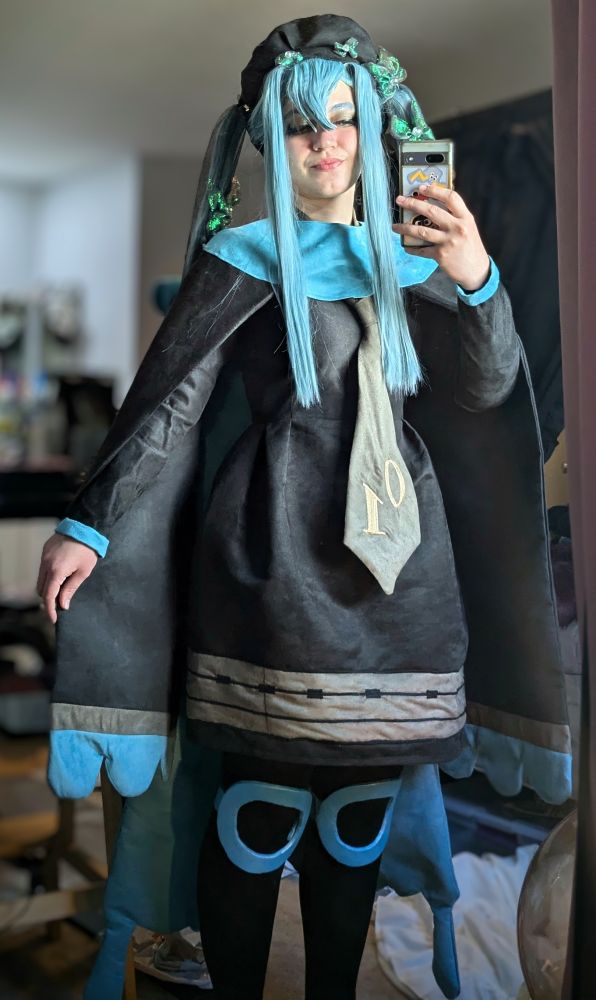 Bug Miku cosplay, selfie pose. Black beret, teal pigtails with green butterflies in the hair, blue collar, black dress, arm capes, tie with 01 on it,  and teal knee pads