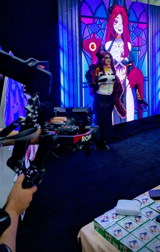 Giwi cosplayer (keybeau) standing next to a large screen with Giwi projected onto it. There is a fancy camera in the foreground 