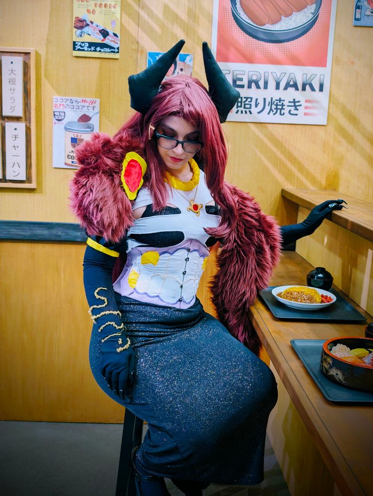 Giwi cosplay, sitting on a stool in front of a ramen bar. Looking downward with a hand on the bar. Food is at the bar. 