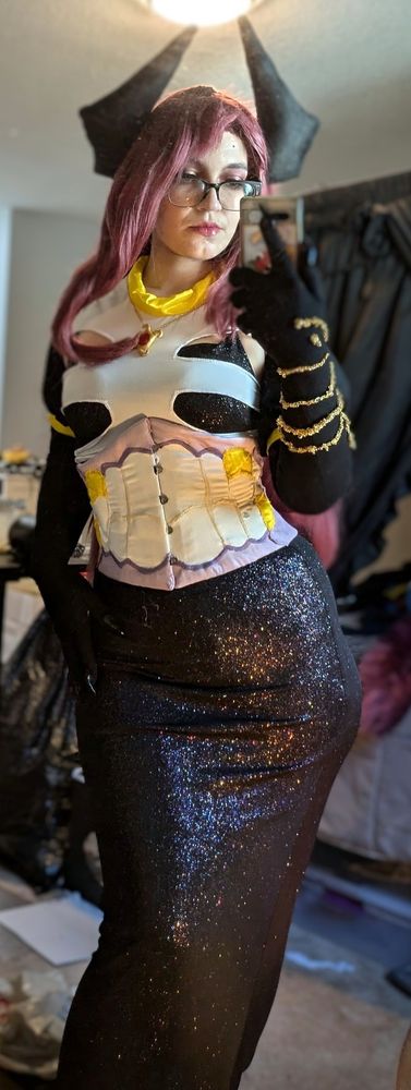 Giwi cosplay, selfie pose. Long maroon wig with pointed black horns, gold pendant, ribcage top, black gloves with gold detailing, teeth-gut corset, sparkly black dress