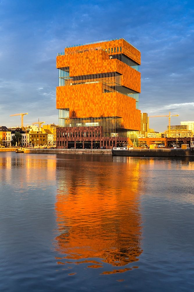An evening view of the 60-metre-high (200 ft) MAS - Museum aan de Stroom - glowing in the light of the setting sun and reflected in the dockside waters of the river Scheldt. The extraordinary building was designed by Neutelings Riedijk Architects; the façades are constructed from Indian red sandstone with curved glass panel infills. It is an example of postmodern Art Deco architecture.