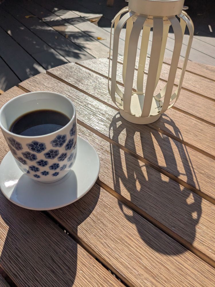 Bright morning sunshine picture of black filter coffee in a blue and white floral motif china cup with no handle, on a wooden table, alongside a candle holder throwing shade across the table.