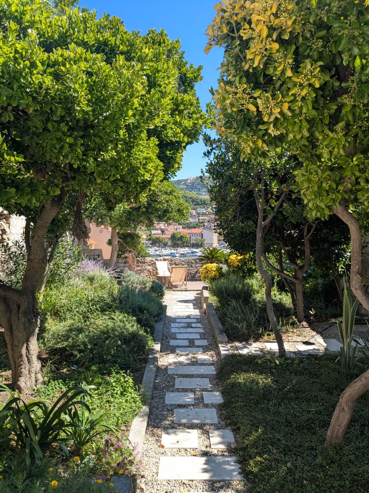 View along a garden path through small trees and Mediterranean herbs, towards a low wall overlooking a sleepy port in a medieval old town. 