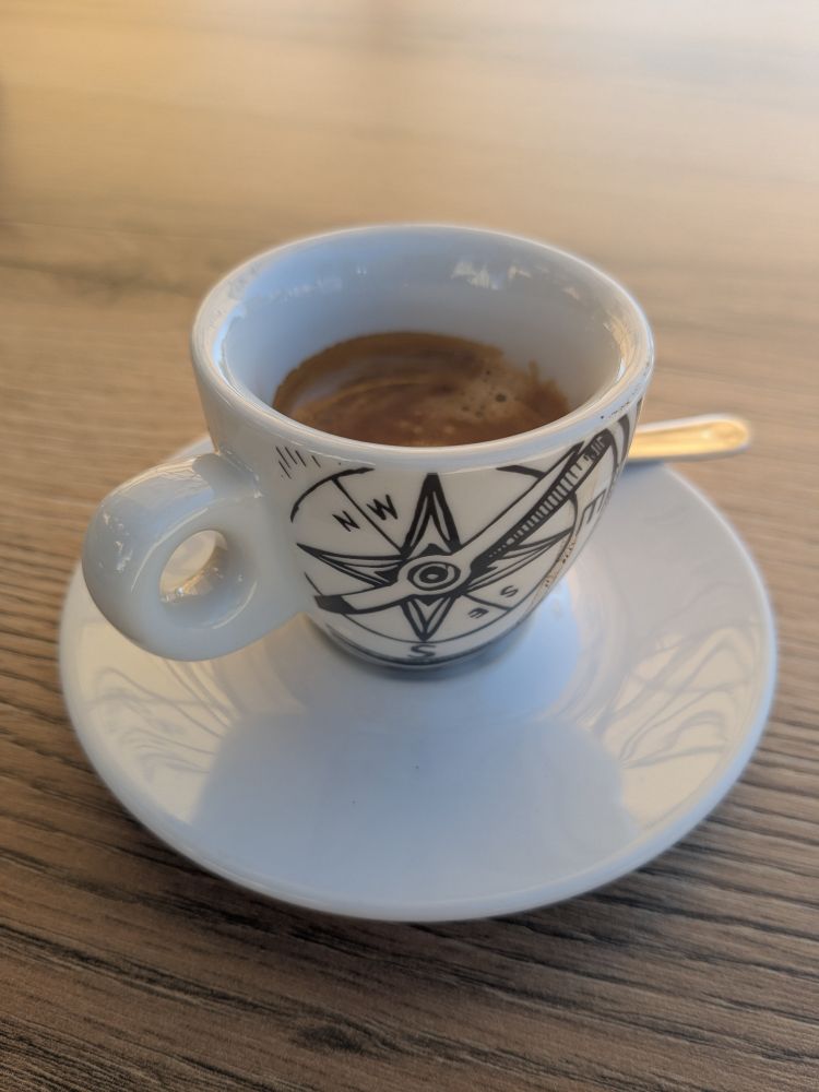 Dark, bitter, over-roaster Mediterranean style old school espresso. In a white cup with a compass motif. 