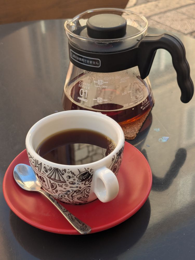 Cup of coffee and a glass hario beaker on a black table. The cup is white and decorated with nautical motifs including octopi. The saucer is orange.