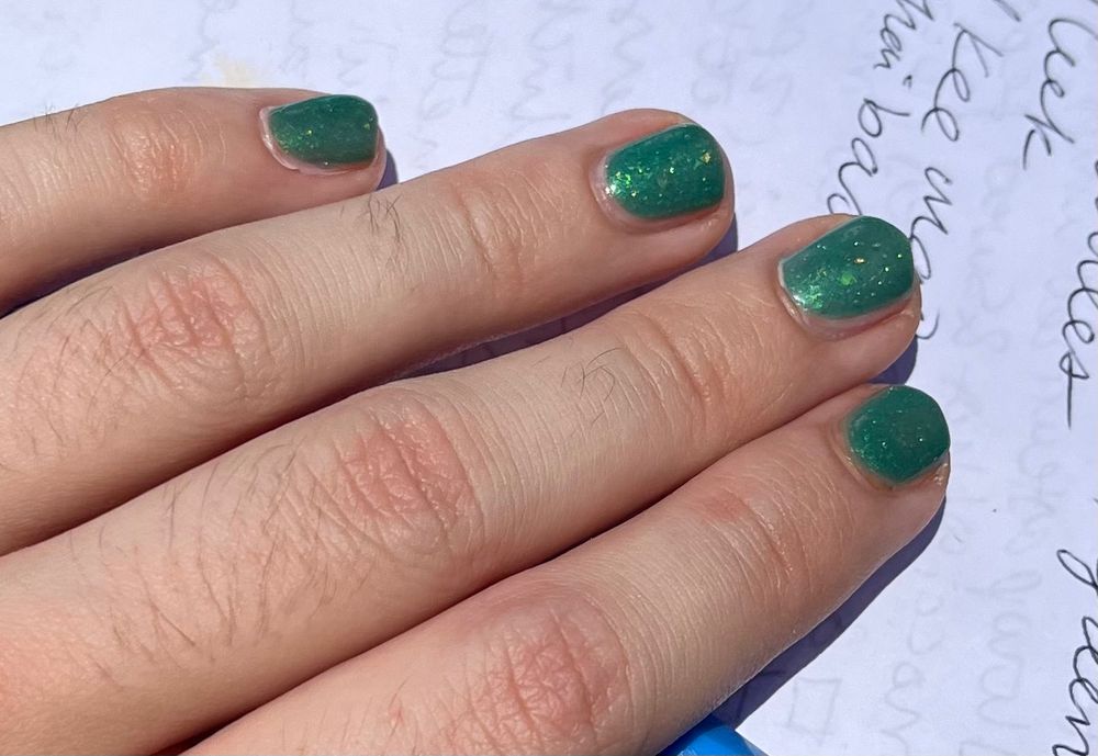 [ID: a cropped photo of a hand with flakie green nail polish on the nails holding a paper with handwriting on it. /end ID.]