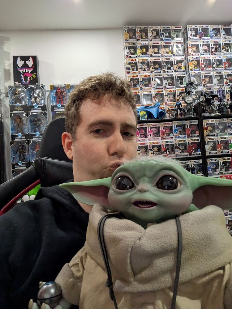 Selfie of me in Daniel's room with many funko pops on the wall. I'm holding up a realistic Grogu toy.