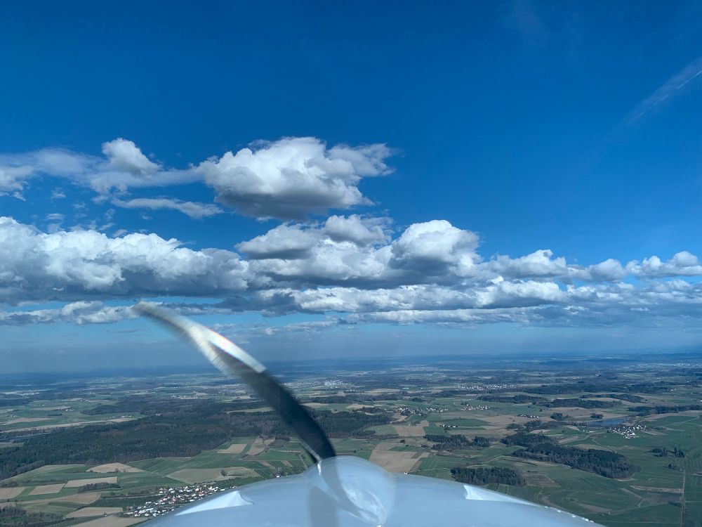 The top of the picture shows blue skies followed by defined cumulus clouds indicating the beginning of the "thermals season". The cowling of an aircraft sneaks into the image from the bottom. 
