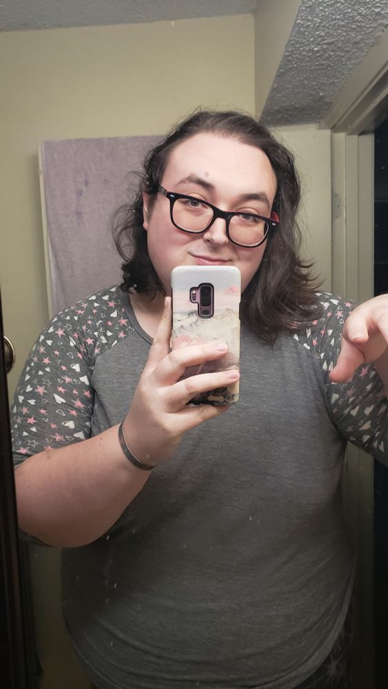 Bathroom mirror selfie of the author, a tall fat white woman in pj's with shoulder length brown hair and black glasses, smiling wanly through the rims