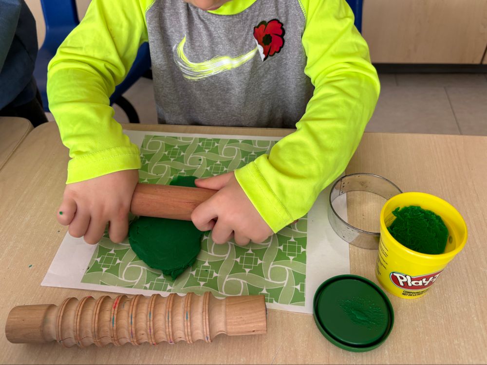A student rolling green play dough makes a smooth pancake.