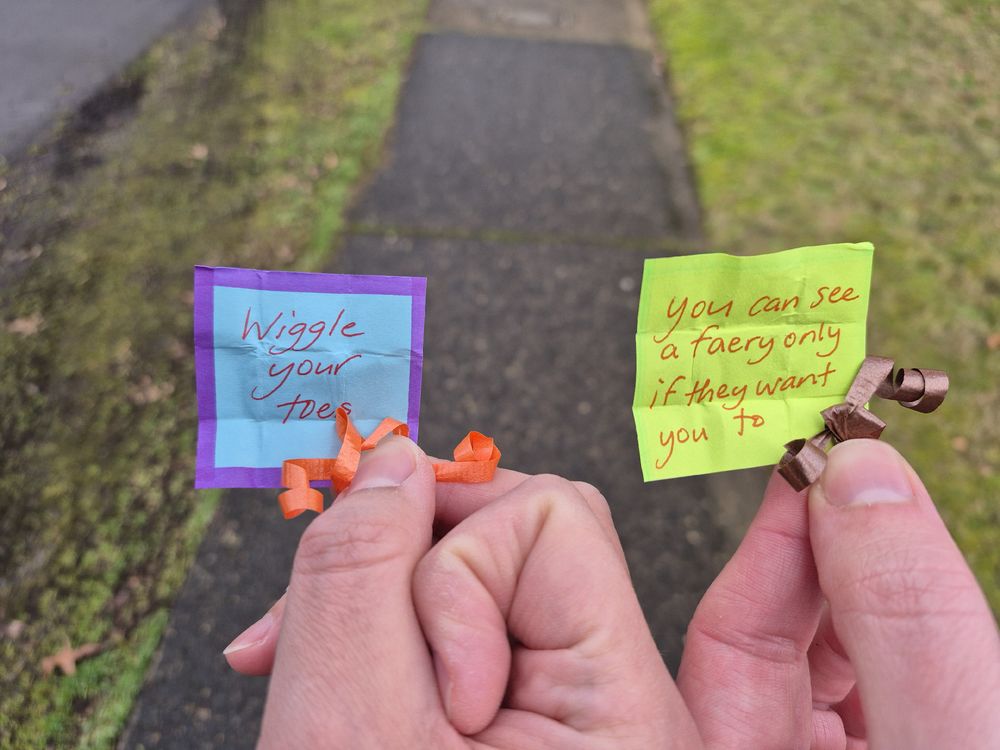 Two hand holding small, brightly colored, hand written notes. The one on the left says "wiggle your toes" and the notes on the right says "You can see a faery only if they want you to". 