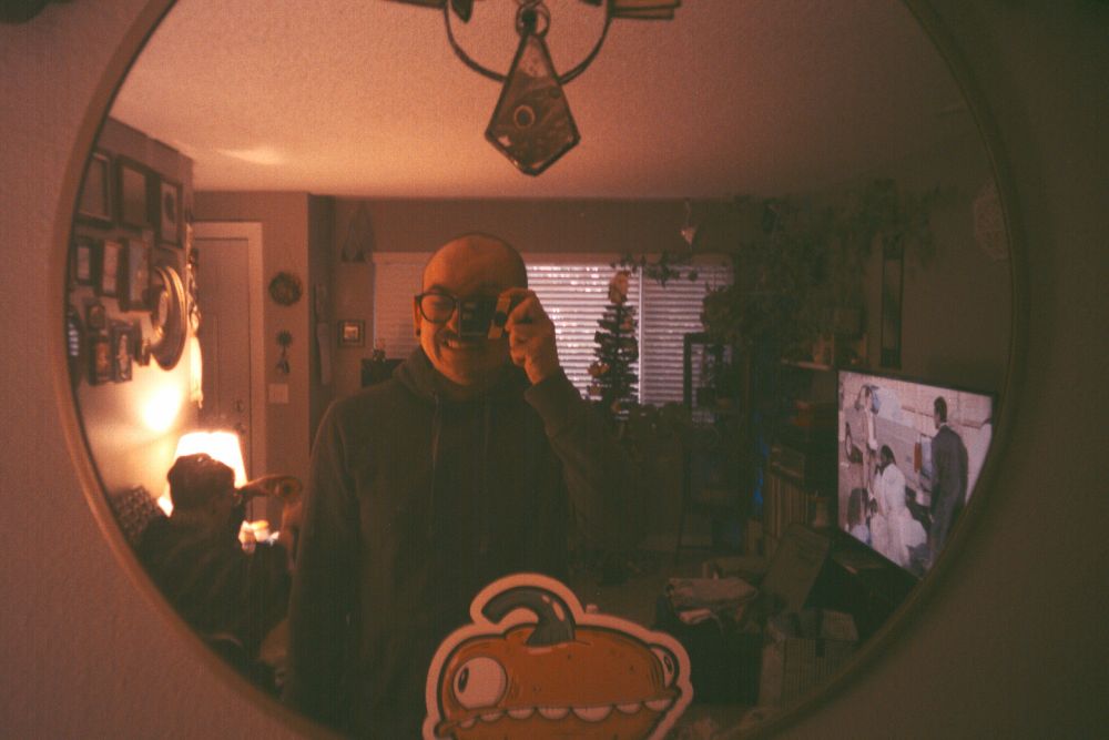 A man takes his photo in the mirror with a small point and shoot camera resembling a film disposable. The room behind him in the mirror shows a Christmas tree, another man sitting on a couch, and a large tv with "The Office" playing on it. There is a decal of a silly pumpkin with a face stuck to the mirror.