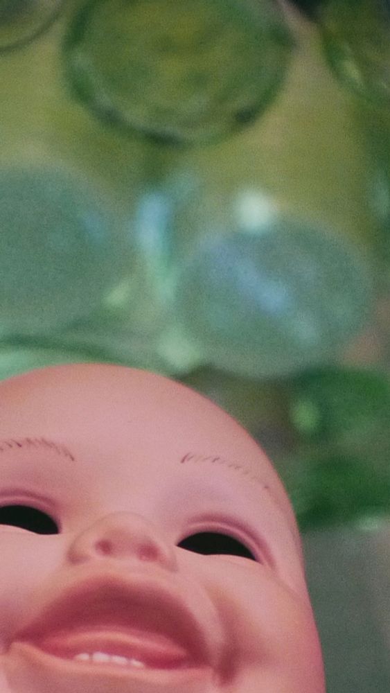 A close crop of an eyeless baby doll head. Bright green glass objects are behind the doll head.