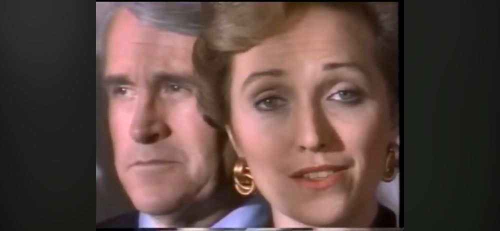 Still from a film or video in which one woman (shot in extreme close up) in formal attire talks to the camera while a man very close behind her stares blankly in space. 