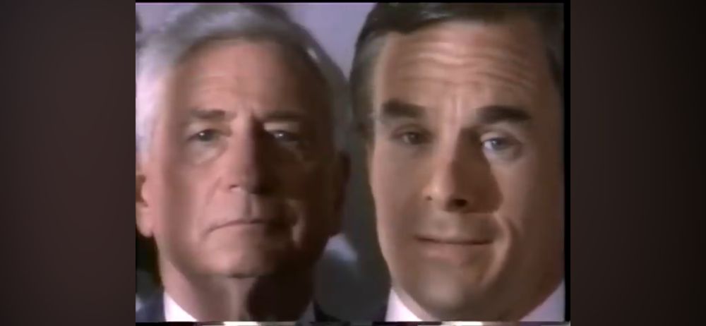 Still from a film or video in which one man(shot in extreme close up) in formal attire talks to the camera while another man very close behind him stares at it. 