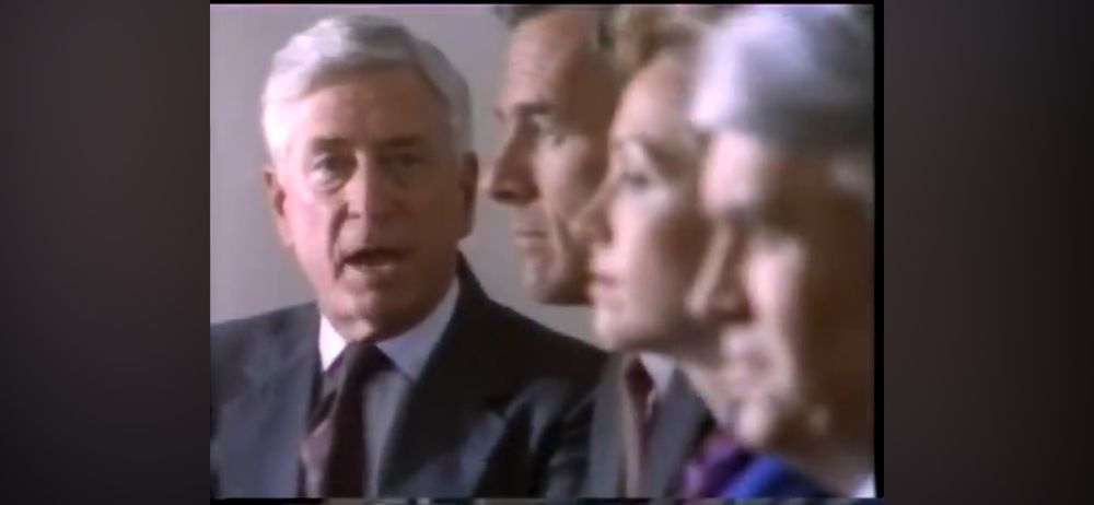 Still from a film or video in which several people (shot in extreme close up) in formal attire stare blankly in space while one person talks to the camera. 