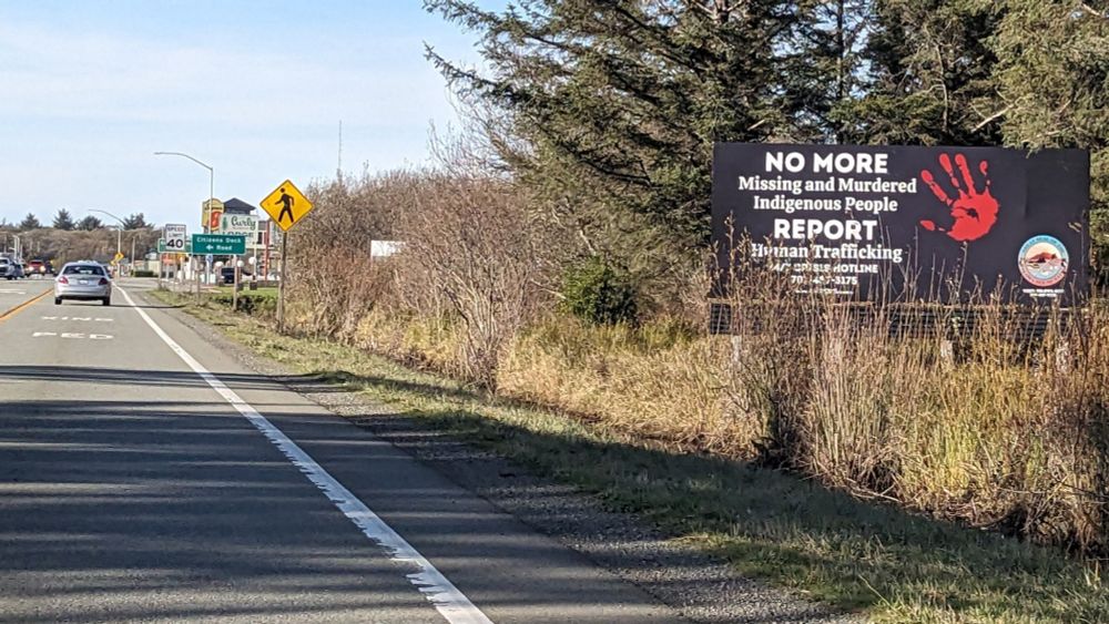 Side of a small highway with a sign with a red paint hand that says "No More Missing and Murdered Indigenous People, Report Human Trafficking" and I can't read the rest