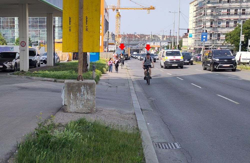 Foto der dreispurigen Richtungsfahrbahn, eine Person radelt auf der Fahrbahn, eine am Gehsteig. Eine Fahrbahn die nach Radweg aussieht, endet vor einer Tankstelle.