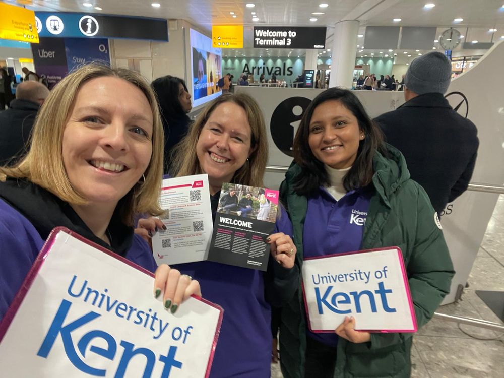Three Kent staff smile while holding up university of kent welcome signs at arrivals in the airport 