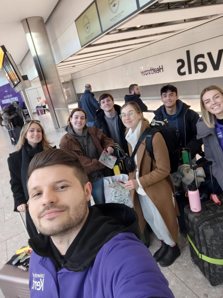 A University of Kent staff member smiles and takes a selfie with our new international students arriving at Heathrow with their luggage 