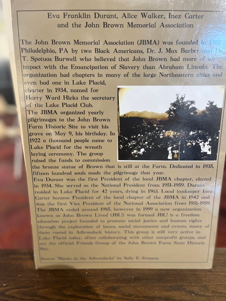 A further exhibit plaque on the John Brown Farm as a public history site which reads: “The John Brown Memorial Association (JBMA) was founded in 1922 [in] Philadelphia, PA by two Black Americans, Dr. J. Max Barber and T. Spotuas Burwell who believed that John Brown had more of an impact with the Emancipation of Slavery than Abraham Lincoln. The organization had chapters in many of the large Northeastern cities and even had one in Lake Placid, charter 1934, named for Harry Ward Hicks the secretary of the Lake Placid Club. The JBMA organized yearly pilgrimages to the John Brown Farm Historic Site to visit his grave on May 9, his birthday. In 1922 a thousand people came to Lake Placid for the wreath laying ceremony. The group raised the funds to commission the Brown statue of Brown that is still at the Farm. Dedicated in 1935, fifteen hundred souls made the pilgrimage that year. Eva Durant was the first President of the JBMA chapter, elected in 1934. She served as the National President from 1951-1959. Durant resided in Lake Placid for 42 years, dying in 1963. Local innkeeper Inez Carter  became President of the local chapter of the JBMA in 1942 and was the first Vice President of the National Association from 1951-1959. The JBMA ended around 1985, however in 1999 a new organization  known as John Brown Lives! (JBL!) was formed. JBL! is a freedom education project founded to promote social justice and human rights through the exploration of issues, social movements and events, many of them rooted in Adirondack history. This group is still very active in Lake Placid today, often collaborating with other nonprofit groups and are the official Friends Group of the John Brown Farm State Historic Site.”