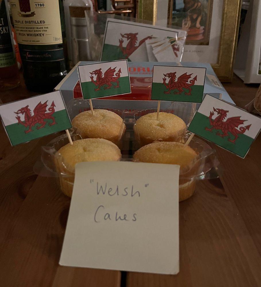 Four Victoria sponge cakes with Welsh flags on cocktail sticks with a sign that says “Welsh” cakes.