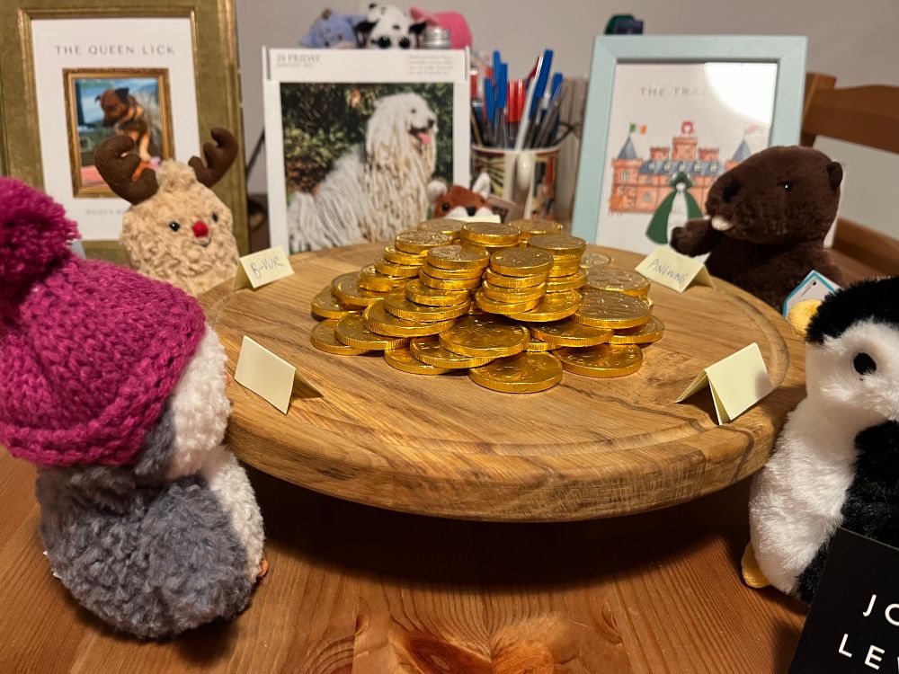 Five soft toy animals sit around a round chopping board, which is piled high with gold chocolate coins 