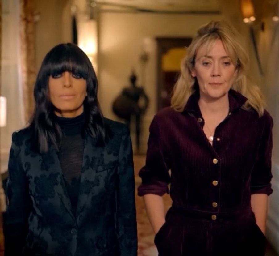 A woman with dark hair and a woman with blonde hair walk down a corridor in a castle looking serious and somber 