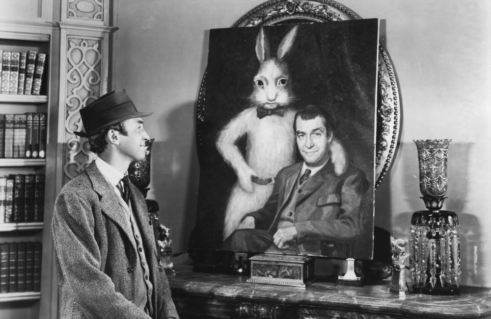 Screencap from the movie Harvey (1950), screenplay Mary Chase. Jimmy Stewart, as Elwood P. Dowd, is smiling at a large painting of Dowd with Harvey. Harvey is a pookah, a tall trickster spirit with a rabbit's head.