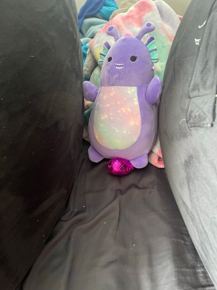 An oval-shaped purple stuffed monster, with antennae, axolotl-like head gills or fins, black dot eyes, and a sharp-toothed smile. The monster's belly is a pastel rainbow dotted with stars.

A pink foil-wrapped egg is sitting at the monster's feet.