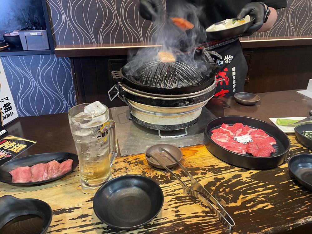 左からラムタン、酎ハイジョッキ、トング、肉の盛り合わせ、アスパラガス
奥の煙が出ているのがジンギスカン用の鉄板
(大黒屋 カウンターテーブル)