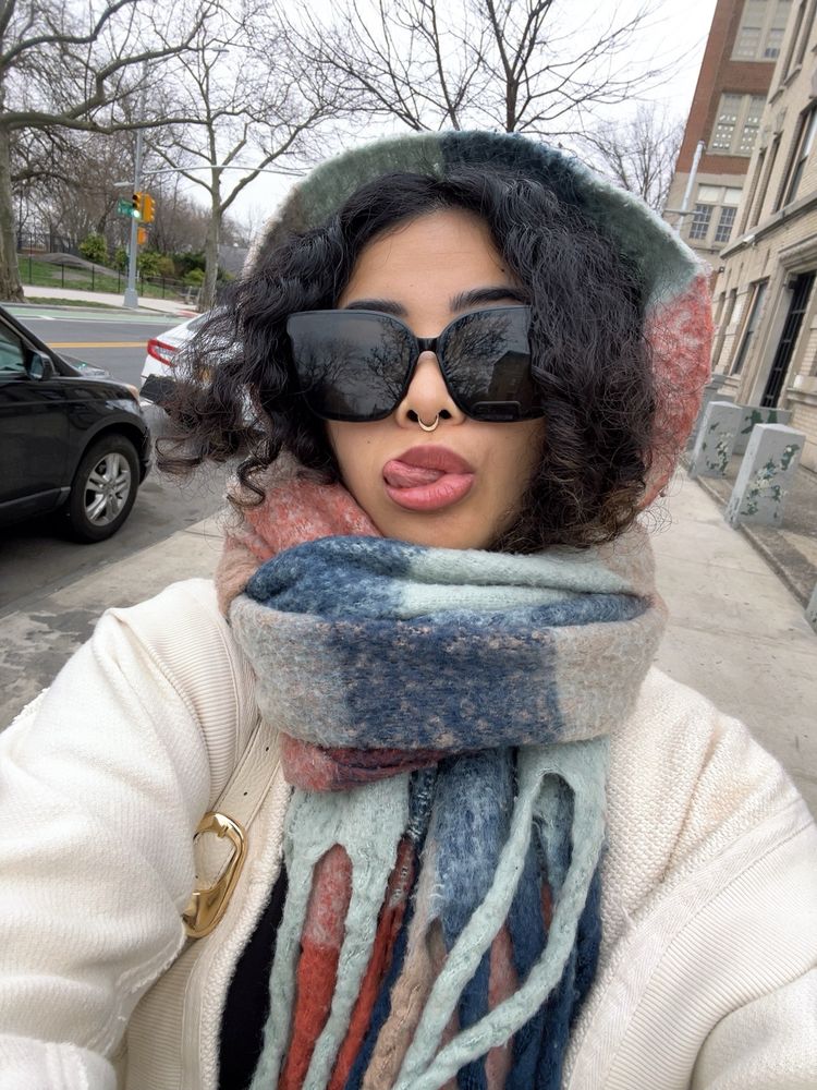 selfie of a person with big black sunglasses, curly hair, and a plaid scarf tied around their head with a white cardigan sticking their tongue out