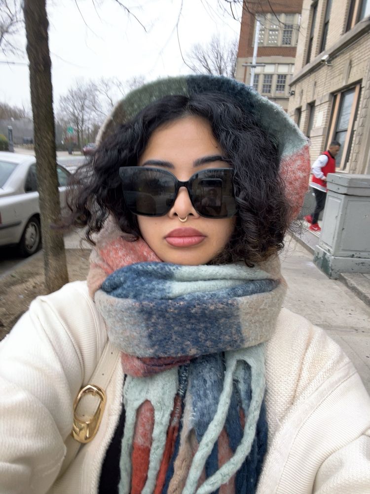 selfie of a person with big black sunglasses, curly hair, and a plaid scarf tied around their head with a white cardigan 