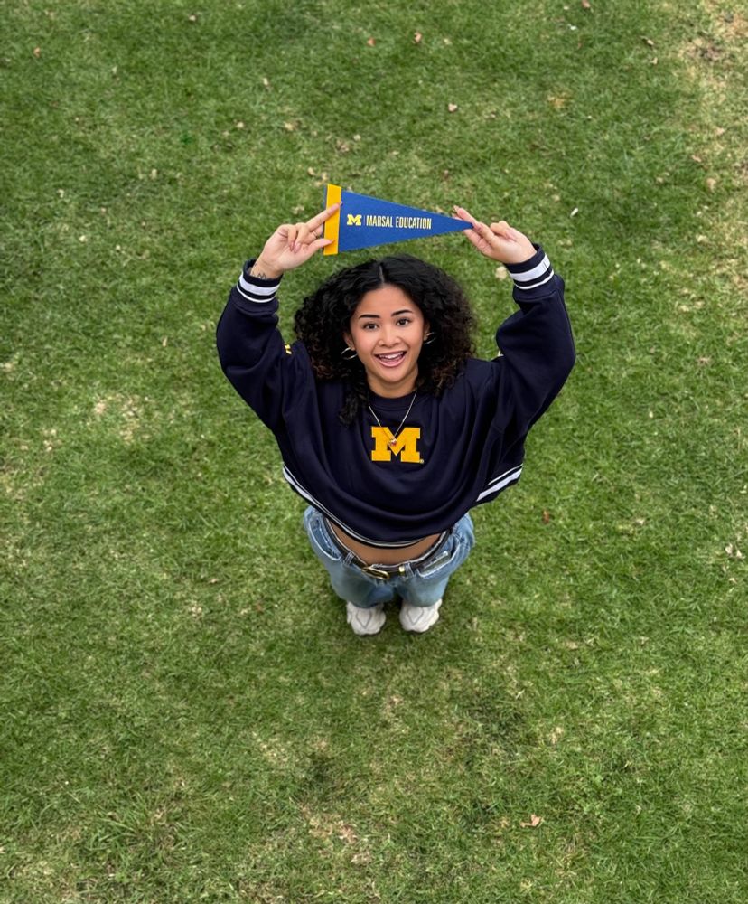 picture of person with curly hair wearing a navy blue University of Michigan crew neck and holding a pennant that says “Marsal Education” above their head. person is standing and looking up at the camera, like an aerial shot.